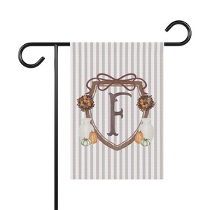 May include: A rectangular garden flag featuring a vertical light gray and white striped pattern. The center displays a brown shield emblem with a large letter F, topped with a bow, flanked by floral arrangements, white vases, and small pumpkins.