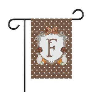 Peut inclure: Un drapeau de jardin rectangulaire avec un fond marron à pois blancs. Au centre, un écusson blanc porte la lettre F, surmonté d'un nœud à carreaux. Deux vases blancs avec du feuillage d'automne et des petites citrouilles entourent l'écusson.