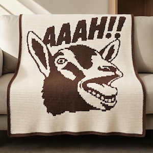 May include: A cream-colored crochet blanket featuring a large, dark brown graphic of a screaming goat head with its mouth open, displaying teeth. Above the goat, the text AAAH!! is written in dark brown, and the blanket has a dark brown border.