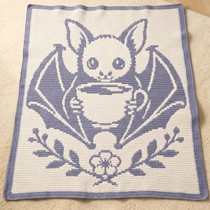 May include: A rectangular crochet blanket featuring a two-tone design in cream and periwinkle blue. The central graphic depicts a bat holding a coffee cup, framed by a floral sprig at the bottom. The blanket is finished with a solid blue border.