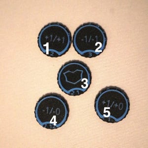 May include: Five circular black tokens with blue borders and textured surfaces. Each token features distinct markings: +1/+1, -1/-1, a central shield-like icon, -1/0, and +1/+0. White numbers 1 through 5 are overlaid on each individual token.