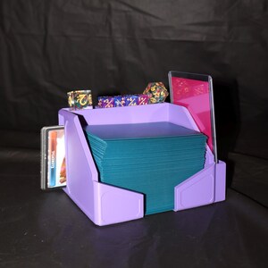 May include: A lavender plastic gaming organizer holding a large stack of teal playing cards. The organizer features side slots for additional cards and a top shelf containing several multi-colored polyhedral dice and a single card in a clear sleeve.
