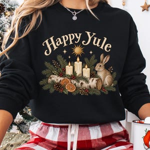 以下が含まれることがあります： Happy Yuleの文字と、3本の灯ったキャンドル、ウサギ、松の枝、丸太に乗ったオレンジのスライスが描かれた黒の長袖スウェットシャツ。赤と白のチェック柄のパンツと、サンタクロースのイラストが入った白いマグカップを合わせています。