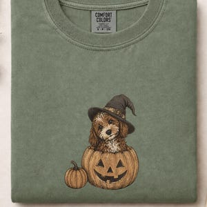 May include: A sage green crewneck t-shirt featuring a graphic of a brown puppy wearing a black witch hat sitting inside a carved jack-o'-lantern pumpkin, with a smaller pumpkin next to it. The inner neck label reads Comfort Colors.
