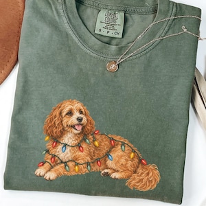 Peut inclure: Un t-shirt à col rond vert olive avec une illustration d'un chien brun doré aux poils bouclés, allongé et entouré d'une guirlande de lumières de Noël colorées. Le t-shirt porte une étiquette de marque Comfort Colors au col avec la taille S.