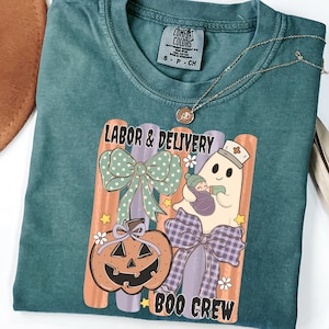 May include: A teal crewneck t-shirt featuring a graphic with the text Labor & Delivery Boo Crew. The design includes a jack-o'-lantern, a ghost wearing a nurse cap holding a swaddled baby, a polka dot bow, and a plaid bow against vertical stripes.