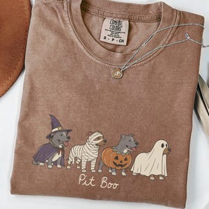 Könnte beinhalten: Ein braunes Kurzarm-T-Shirt mit einer Grafik von vier Hunden in Halloween-Kostümen: eine Hexe, eine Mumie, ein Kürbis und ein Geist. Unter den Hunden steht der Text Pit Boo. Das Shirt hat ein Comfort Colors Etikett am Hals und eine silberne Halskette.