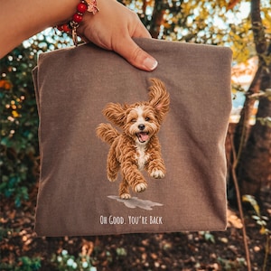 Könnte beinhalten: Ein braunes T-Shirt mit einer Grafik eines fröhlichen, rennenden, lockigen braun-weißen Hundes. Unter dem Hund steht der Text "OH GOOD. YOU'RE BACK.". Der Hund hat die Zunge herausgestreckt, die Ohren wehen im Wind und er steht über einer grauen Pfütze.