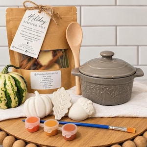 May include: A holiday simmer pot kit featuring a brown paper pouch with a tag, a wooden spoon, a gray ceramic pot with a lid, a small striped pumpkin, three white ceramic shapes including a pumpkin, leaf, and acorn, and a paint set with a brush.