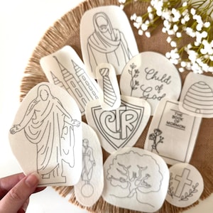 May include: A collection of white fabric patches featuring black line art illustrations including a figure with outstretched arms, a temple, a tie, a shield with CTR text, a beehive, a tree, a cross, and a book labeled The Book of Mormon.