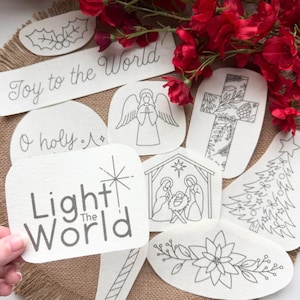 May include: A collection of white fabric patches featuring black line art designs including a nativity scene, a cross, an angel, a Christmas tree, a poinsettia, and text reading Joy to the World and Light The World.