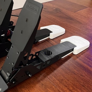 May include: A black metal racing pedal set featuring three upright pedals with vertical plates. The two rightmost pedals are equipped with white plastic pedal covers. The base unit is constructed from dark metal with visible screw holes.