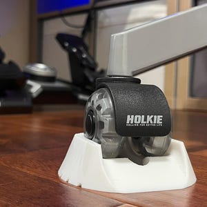 May include: A grey chair caster wheel with a black cover featuring the white text HOLKIE and the slogan ROLLING FOR BETTER LIFE, secured within a white plastic floor cup stabiliser. The wheel is attached to a grey metal chair leg frame.