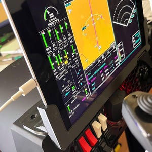 May include: A tablet mounted in a black plastic stand displaying a flight simulation interface with various gauges, engine data, and navigation maps. A white charging cable is plugged into the side of the tablet, which is positioned next to a flight controller with red and white toggle switches.