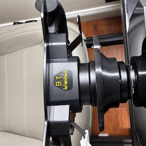 May include: A black Moza Racing steering wheel base with a central yellow label displaying the number 16. The unit features a metallic quick-release mechanism and a black mounting plate with the Moza logo printed in white on the side.