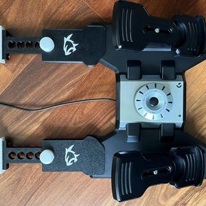 Puede incluir: Un conjunto de pedales de juego en negro y plata que incluye dos pedales texturizados, un dial circular central plateado con el logotipo de Logitech G y dos brazos negros ajustables con iconos de aleta de tiburón y perillas blancas.