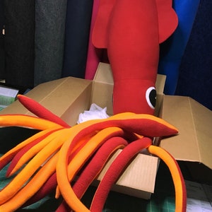 Puede incluir: Un calamar de peluche grande de color rojo y naranja con tentáculos en una caja de cartón. El calamar tiene una cabeza grande y un cuerpo largo con un solo ojo grande.