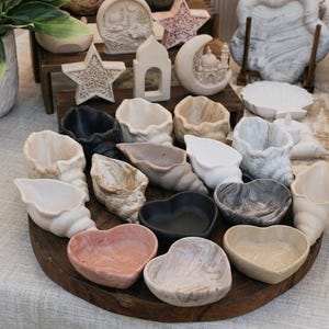 May include: A collection of small decorative bowls and ornaments made of resin or stone. The items include heart-shaped dishes in pink, beige, and black, shell-shaped containers in various marbled tones, and sculpted figures of stars, moons, and arches.