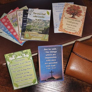 Puede incluir: Una colección de tarjetas rectangulares con versículos bíblicos impresos sobre fondos de naturaleza, incluyendo Filipenses 2:13, Salmo 56:3, 2 Corintios 5:17 y Lucas 18:27, exhibidas junto a un libro encuadernado en cuero marrón y una cartera.
