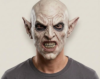May include: A full-head latex mask featuring a pale, wrinkled creature with pointed ears, deep-set dark eyes, and a snarling expression revealing sharp, yellowed teeth. The mask has a textured, weathered skin appearance and is worn over a gray t-shirt.