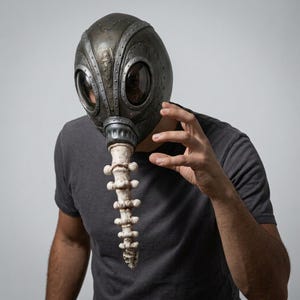 May include: A dark metallic gray full-face mask featuring large circular eye openings with dark lenses, decorative rivets along the seams, and a long, white, segmented bone-like protrusion extending downward from the center of the face.