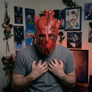 Peut inclure: Un masque intégral en forme de cœur humain réaliste, de couleur rouge vif avec des détails de veines sombres en relief. Le masque présente des découpes pour les yeux et le nez, couvrant tout le visage et la tête avec un design anatomique texturé.