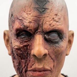 May include: A full-head mask featuring a textured, weathered skin appearance with deep wrinkles and scarring. The right side displays dark, reddish-brown tones, while the left eye is clouded and white. A black triangular symbol is centered on the forehead.
