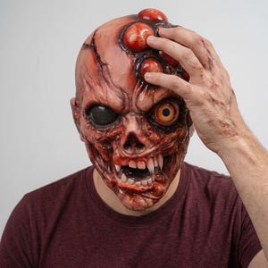 May include: A full-head latex mask featuring a grotesque, fleshy, and textured surface with multiple protruding red bumps on the forehead. The mask displays one solid black eye and one realistic orange eye, along with sharp, pointed white teeth.