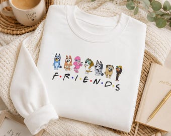 Könnte beinhalten: Ein weißes Langarm-Sweatshirt mit einer Grafik von sieben Zeichentrickhunden, die in einer Reihe über dem Schriftzug F R I E N D S stehen, wobei sich zwischen den Buchstaben bunte Punkte befinden. Das Design zeigt verschiedene Hunderassen.