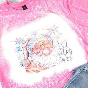 Könnte beinhalten: Ein leuchtend pinkes, gebleichtes T-Shirt mit einer Grafik vom Weihnachtsmann, der eine Discokugel-Mütze und eine Brille trägt, eine Kaugummiblase bläst und ein Friedenszeichen zeigt, umgeben von bunten, aquarellartigen Schneeflocken.
