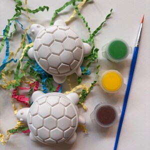 May include: Two white turtle-shaped figurines with a hexagonal shell pattern, accompanied by three small circular paint pots containing green, yellow, and brown paint, and a single thin paintbrush with a blue handle and orange bristles.