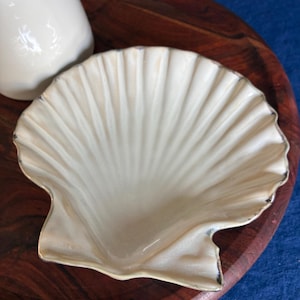 Puede incluir: Un plato de cerámica color crema con forma de concha de vieira, acabado brillante y un sutil borde plateado metálico en los extremos festoneados. El plato presenta estrías radiantes que se extienden desde la base hasta el borde.