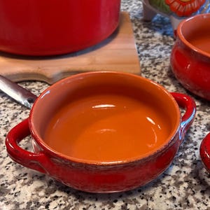 May include: A round, shallow ceramic bowl with two side handles, featuring a vibrant red exterior glaze and a smooth, unglazed terracotta-colored interior. The bowl has a rustic, textured finish along the outer rim and base.