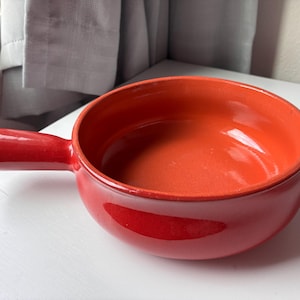 May include: A round, glossy, bright orange ceramic bowl with a single short, sturdy handle extending from one side. The interior and exterior feature a smooth, uniform finish with a deep, vibrant orange color throughout the entire piece.