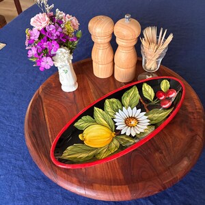 Könnte beinhalten: Ein rundes Holztablett mit einer ovalen schwarzen Keramikschale, die mit einer gelben Zitrone, einer weißen Margerite und roten Beeren bemalt ist. Daneben stehen zwei helle Holz-Salz- und Pfeffermühlen, ein Glas mit Zahnstochern und eine kleine Vase.