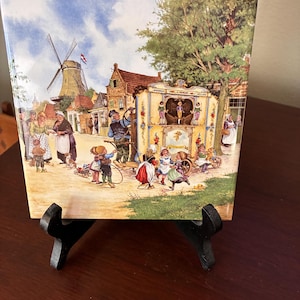 May include: A square ceramic tile featuring a colorful illustration of a Dutch village scene with a windmill, a street organ with the text C. PERLEE, and people in traditional clothing. The tile is displayed on a black plastic stand.