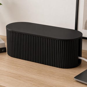 May include: A matte black cable management box with an oval shape and a vertical ribbed texture on the exterior. The container features a solid flat lid and a side opening that allows a white power cord to pass through the side of the enclosure.