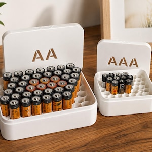 May include: Two white plastic battery storage cases with hinged lids. The larger case is labeled AA and contains multiple orange and black AA batteries. The smaller case is labeled AAA and holds several smaller black and orange AAA batteries.