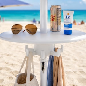 May include: A white circular beach umbrella table with a central pole, featuring four hooks holding a beige tote bag, a set of keys, a blue towel, and a tan towel. On the tabletop sit brown aviator sunglasses, a soda can, and a tube of SPF 50 sunscreen.