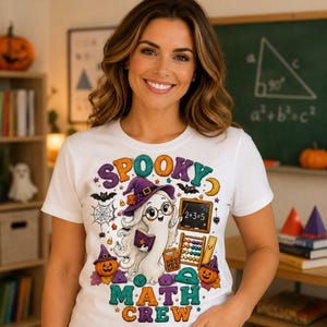 May include: A white t-shirt featuring a colourful graphic of a ghost wearing a witch hat and glasses, holding a book. The design includes a chalkboard with 2+3=5, an abacus, a calculator, pumpkins, bats, and the text SPOOKY MATH CREW.