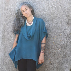 Op de afbeelding: Een vrouw met grijs haar draagt een tealblauwe top met een cowl-hals en een witte kralenketting. Ze heeft een zwarte tatoeage op haar arm.