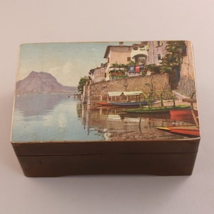 May include: A small, rectangular wooden box with a painted landscape scene on the lid. The artwork depicts a lakeside village with boats, buildings, and a mountain in the background. The box is brown.