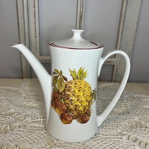 May include: A white porcelain coffee pot featuring a central illustration of a yellow pineapple surrounded by smaller brown fruits and green leaves. The pot has a matching lid with a knob, a curved handle, a long spout, and a thin red rim accent.