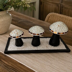 May include: Three mushroom-shaped decorative figures with black stems and mosaic-tiled caps in white and iridescent tones. The mushrooms are arranged in a row on a rectangular black tray filled with small white pebbles as a base for the display.