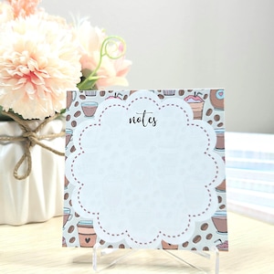 May include: A square notepad with a scalloped circular center featuring the word notes in black script. The border displays a pattern of coffee cups and beans in shades of brown, pink, and blue on a light gray background.