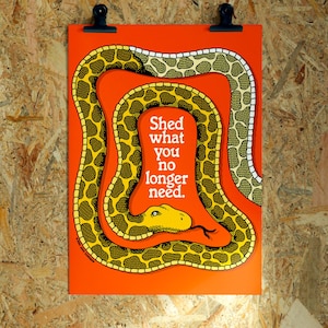 Könnte beinhalten: Ein rechteckiges orangefarbenes Poster mit einer geringelten, gelb-schwarz gemusterten Schlange, deren Kopf und Zunge sichtbar sind. In der Mitte der Schlangenspirale steht der weiße Text: Shed what you no longer need. Das Poster wird von zwei schwarzen Clips gehalten.