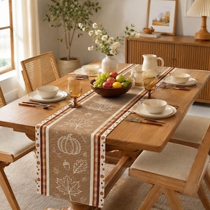 Pode incluir: Um caminho de mesa de tecido castanho com desenhos de linhas brancas de abóboras, bolotas e folhas de ácer sobre uma mesa de madeira. O caminho tem bordas às riscas e está posto com pratos, taças de cerâmica branca e fruta fresca.