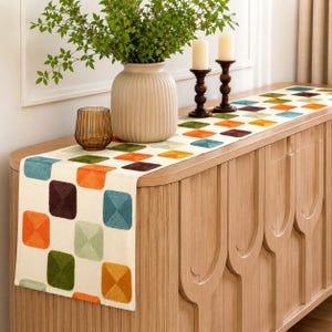 May include: A cream-colored table runner featuring a repeating pattern of squares in shades of orange, teal, olive green, and dark brown. The runner is laid flat on a light wood sideboard, accompanied by a ribbed vase, a glass tumbler, and two candles.