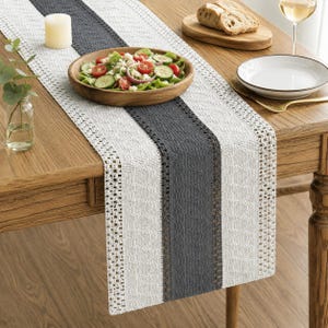 May include: A rectangular table runner featuring a central dark gray textured panel flanked by white woven sections with intricate openwork lace borders. The runner is laid flat on a wooden table, providing a base for a wooden salad bowl.