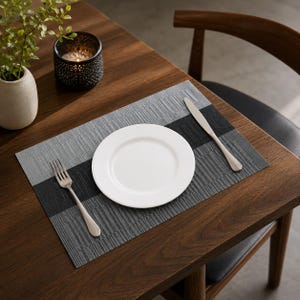 Puede incluir: Un mantel individual rectangular con un diseño texturizado de tres colores con franjas gris claro, negro y gris oscuro. Centrado en el mantel hay un plato de cerámica blanco liso, acompañado por un tenedor de acero inoxidable a la izquierda y un cuchillo a la derecha.